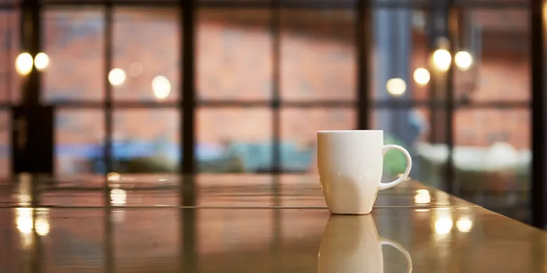 White ceramic coffee cup on a reflective wooden table in front of softly blurred window panes